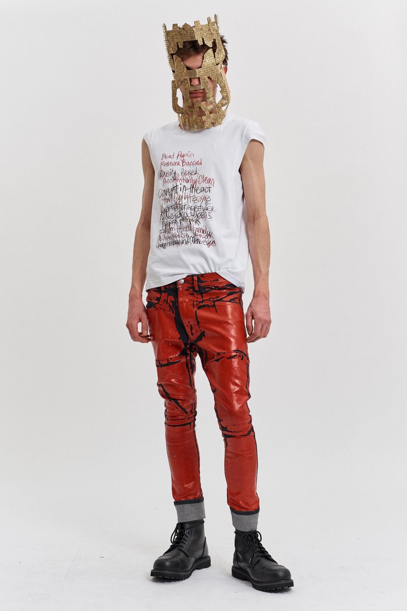 A full shot of a person in statement red pants, black boots, and a mask, posing against a white backdrop in a studio setting.