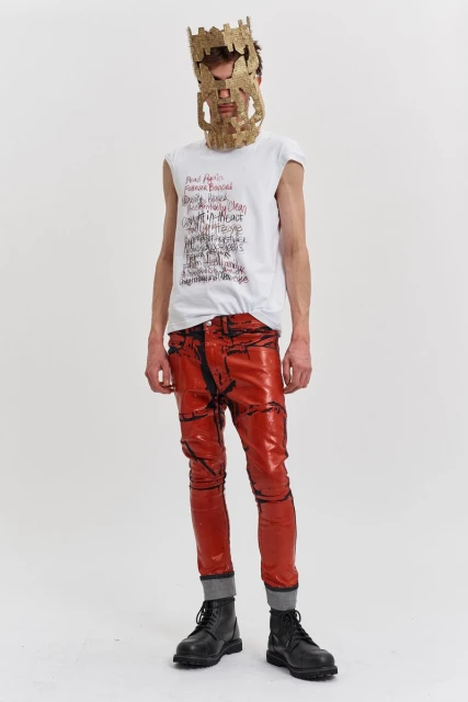 A full shot of a person in statement red pants, black boots, and a mask, posing against a white backdrop in a studio setting.
