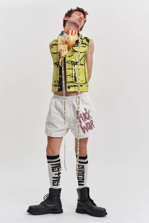 Male fashion model poses in a yellow printed vest, white shorts with writing, black boots, and high socks against a white backdrop.