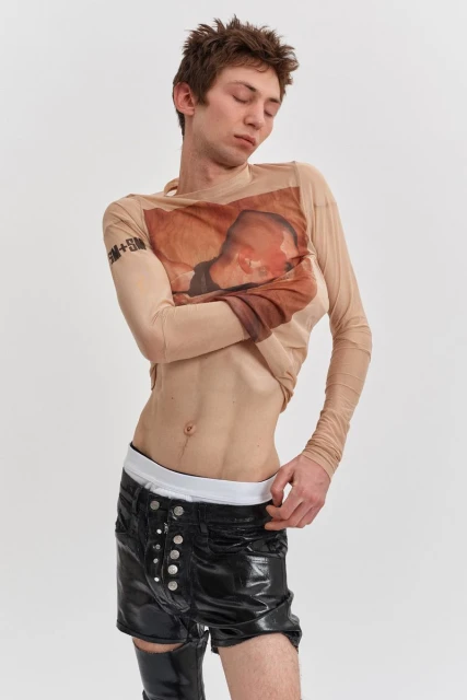 An androgynous person poses in a beige long-sleeved shirt and black shorts with eye contact, against a white backdrop in a studio setting.