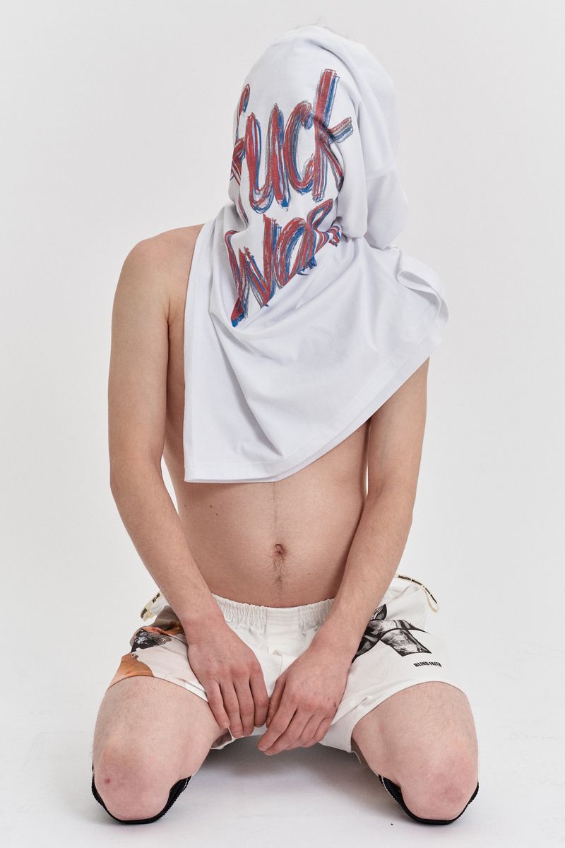 Man kneeling, face covered by white t-shirt with bold blue and red text, knee pads, and printed shorts against a plain white backdrop.