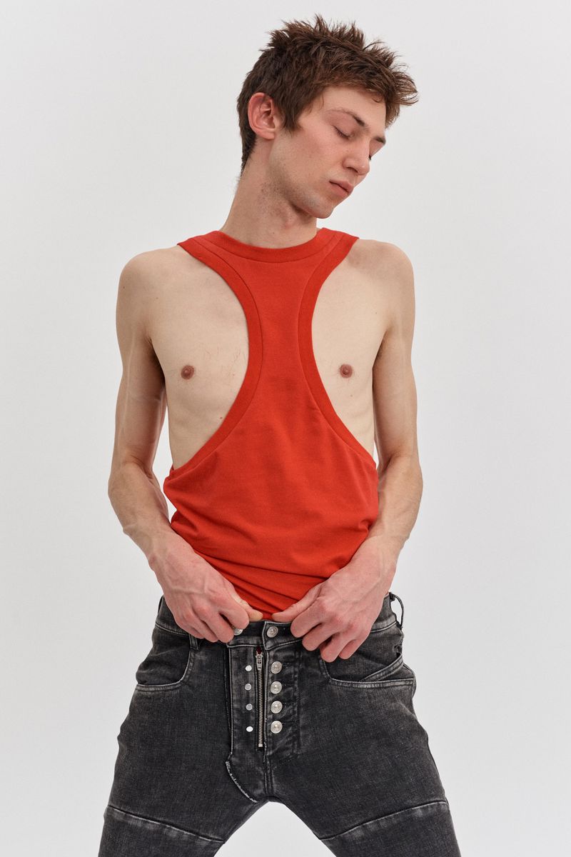 A slender man poses in a red tank top and black jeans against a white backdrop, head tilted down, embodying a fashion-forward, minimalist aesthetic.