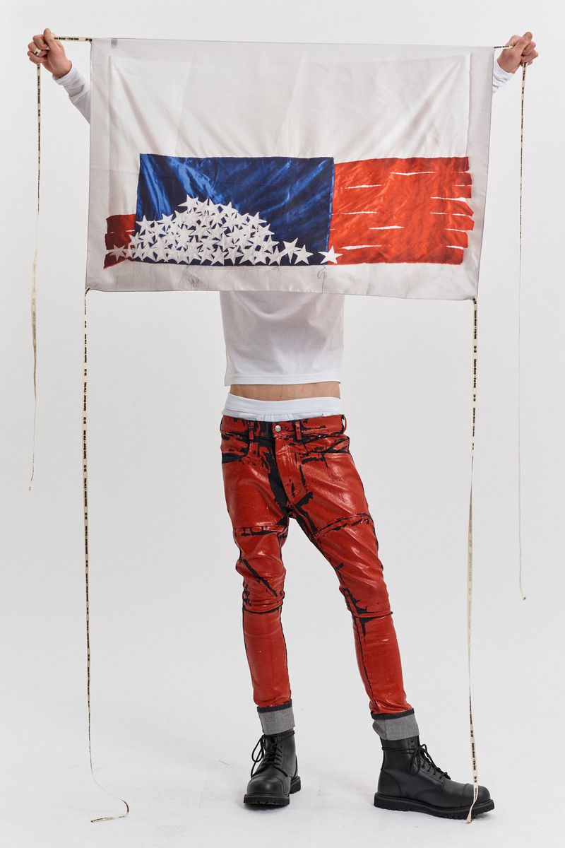 Person holding a flag-inspired textile art piece with red patent leather pants and black boots against a solid white backdrop.