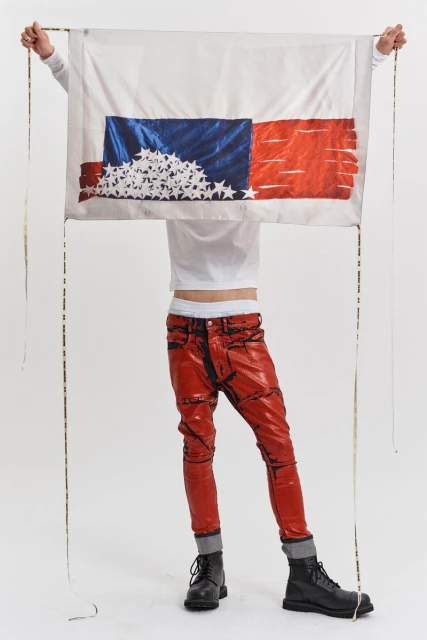 Person holding a flag-inspired textile art piece with red patent leather pants and black boots against a solid white backdrop.