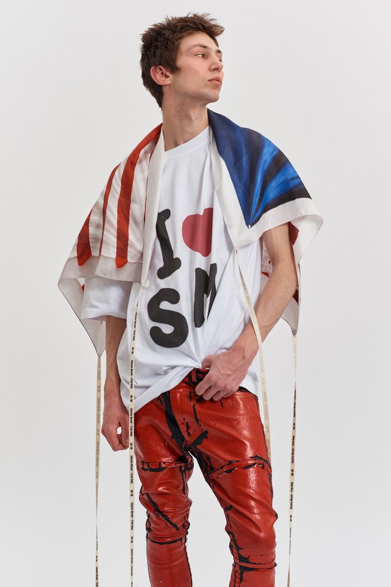 Male model poses in a white t-shirt, red pants, and flag-themed scarf in a bright studio setting for a fashion editorial shoot.