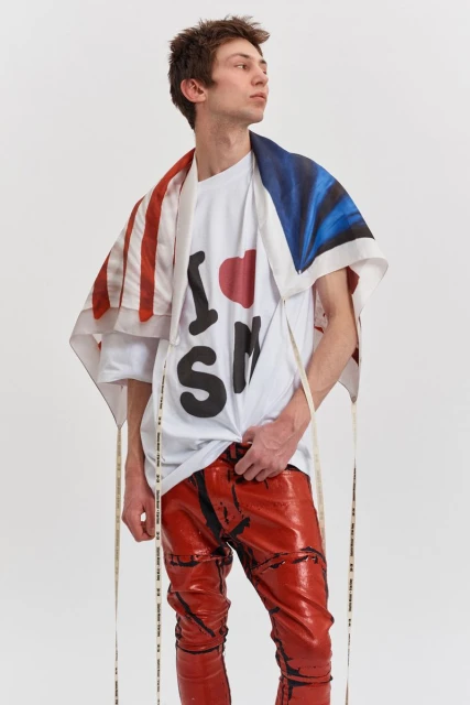 Male model poses in a white t-shirt, red pants, and flag-themed scarf in a bright studio setting for a fashion editorial shoot.
