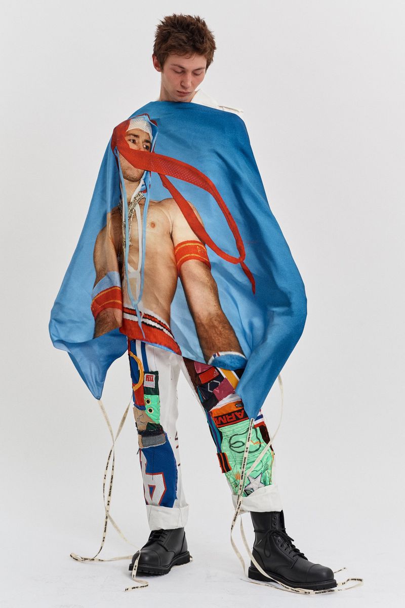 Young man standing indoors wearing graphic pants, black boots, and a blue cape-like garment with a printed image. Fashion editorial.