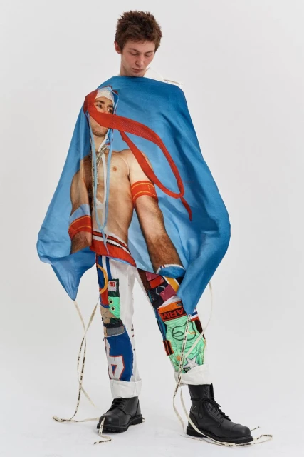 Young man standing indoors wearing graphic pants, black boots, and a blue cape-like garment with a printed image. Fashion editorial.