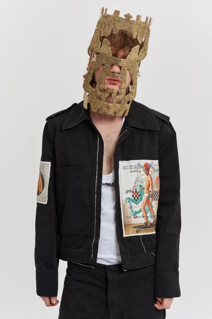 Man in black jacket and gold mask with patches on jacket standing against white background, studio lighting.