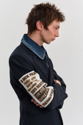 Model in profile wearing a dark tailored blazer with a denim collar and a printed text arm patch against a neutral studio backdrop.