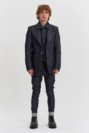 A male model standing in a dark raw denim suit and black lace-up boots against a seamless white studio background.