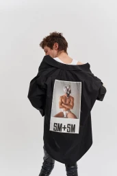 Model seen from the back wearing an oversized black graphic shirt off the shoulder against a plain white studio background.