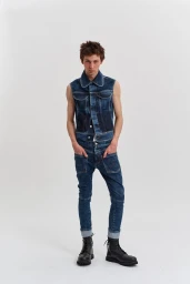 Young male model wearing matching dark denim vest and jeans with black boots against a plain white studio background.