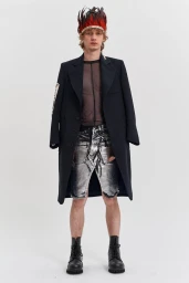 Model wearing dark coat, sheer top, and silver distressed shorts with feathered headpiece on white seamless background