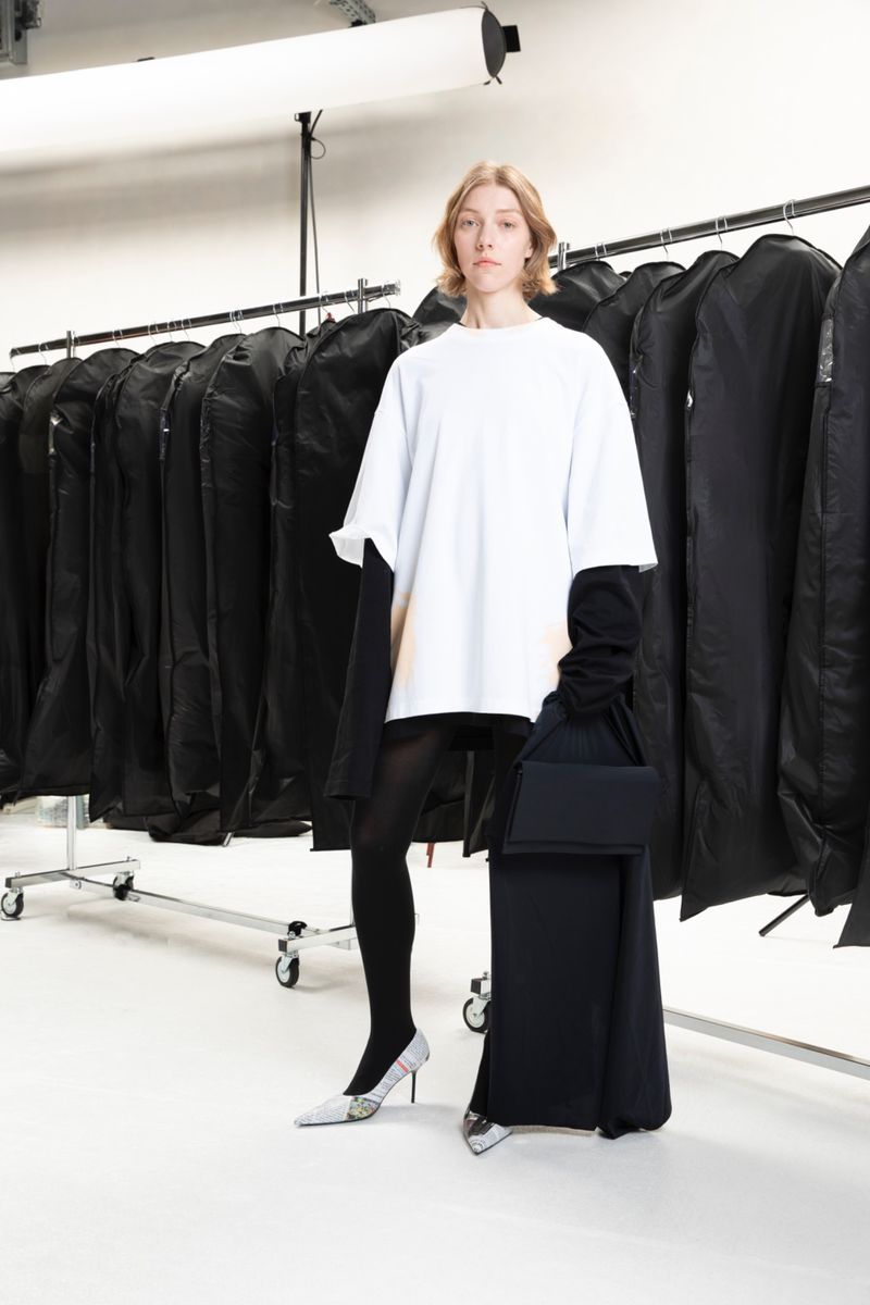 Fashion shot of a woman wearing a white t-shirt, black tights, and silver heels, standing in front of racks of black garment bags in a studio.