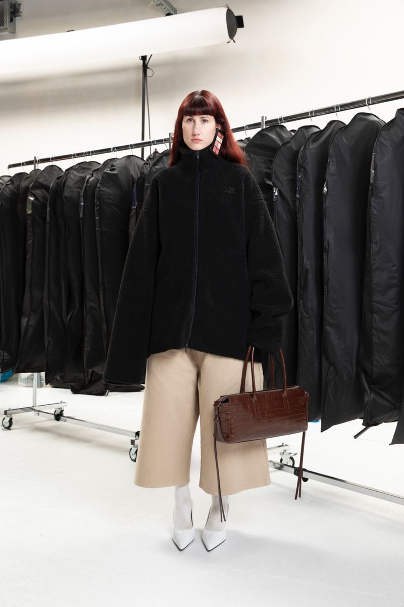 A female fashion model poses in a studio wearing a black fleece jacket, beige culottes, white boots, and carrying a brown leather bag.