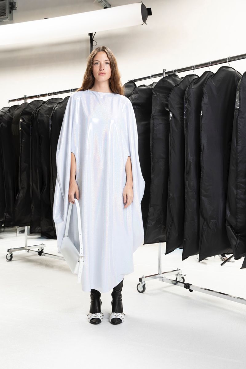Fashion shot of a woman in a white dress holding a white purse, standing in a photo studio with a row of garment bags.