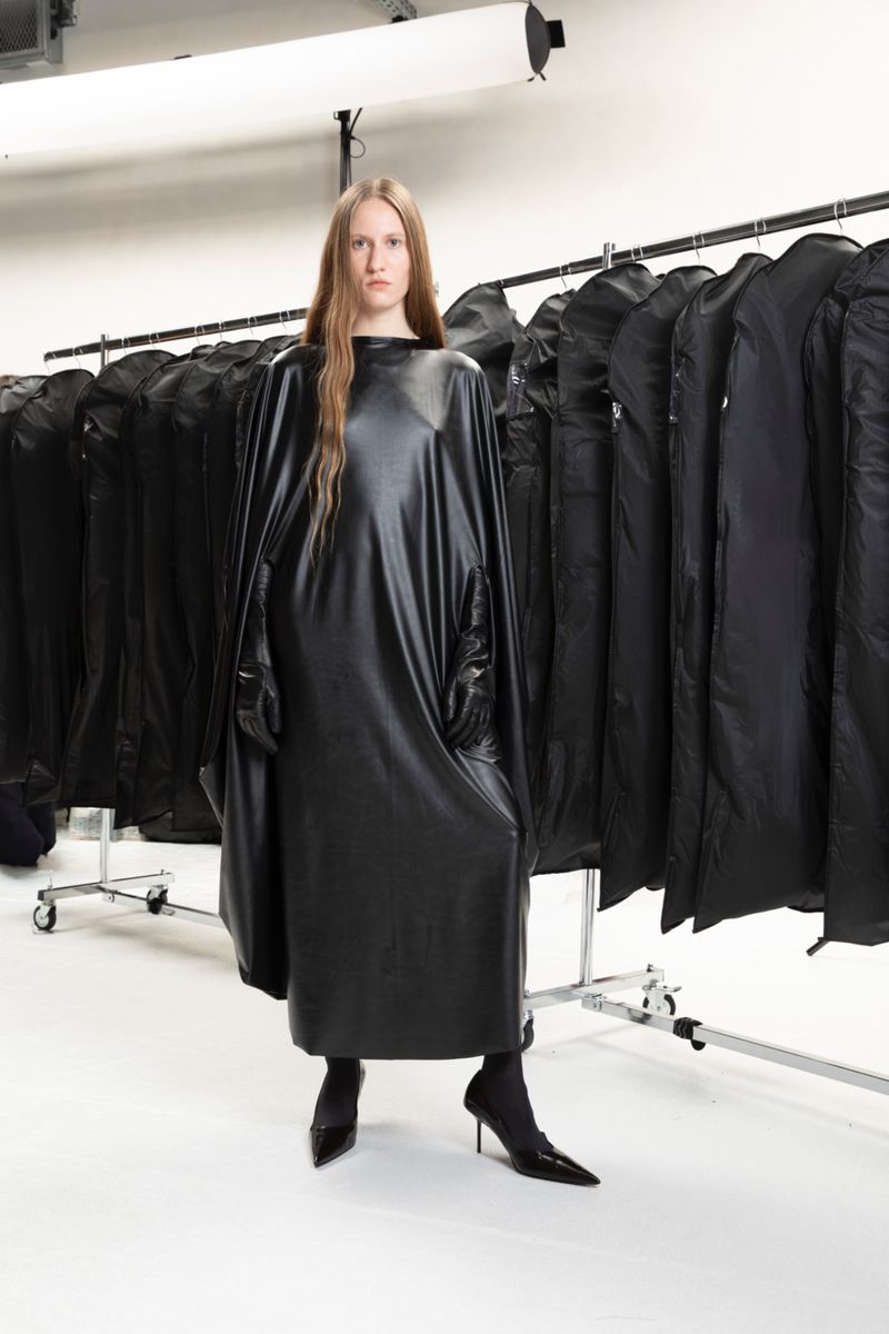 A slender fashion model poses indoors wearing a long, draped, black dress and matching heels, showcasing a high-fashion, modern aesthetic.