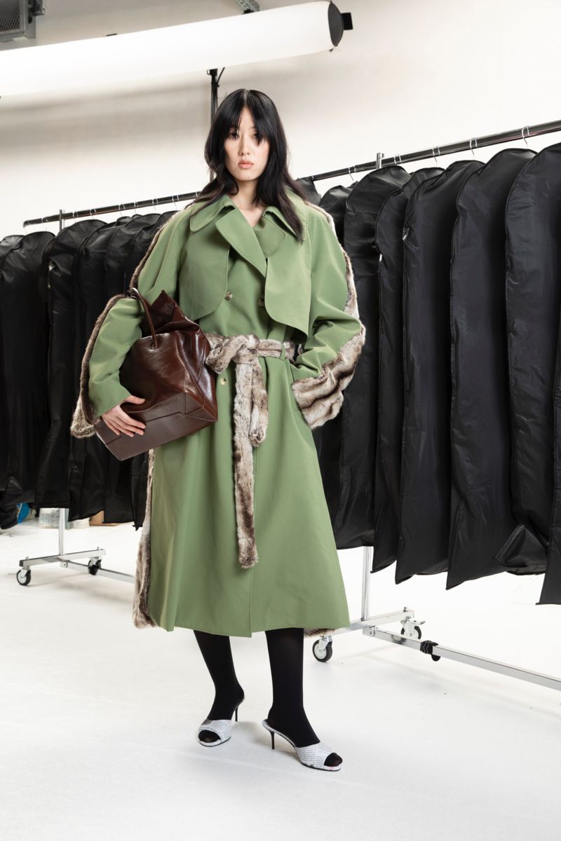 Asian fashion model poses in a green trench coat with fur trim, black stockings, and white shoes against a background of black garment bags.