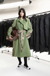 Model wearing a long green trench coat and holding a brown leather bag in front of a clothing rack inside a bright white studio.
