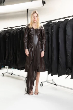 Model standing in a brown leather fringe dress against a white studio seamless backdrop with clothing racks.