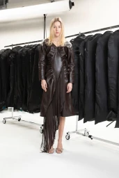 Model standing in a brown leather fringe dress against a white studio seamless backdrop with clothing racks.
