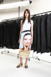 Model standing in a white studio wearing a graphic printed midi dress and brown sneakers against a clothing rack