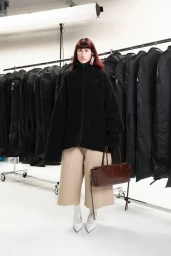 Model wearing a black fleece jacket and beige culottes holding a brown handbag in a studio with clothing racks.