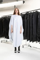 Model wearing a white cape dress and carrying a white handbag in a studio with clothing racks