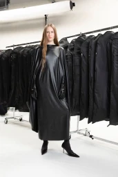 Model standing in a glossy black cape dress in front of a clothing rack inside a bright white studio setting.