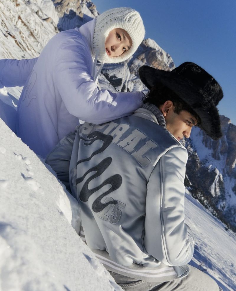 Two fashion models pose in a snowy mountain setting, one in a white hooded jacket, the other in a jacket with a black furry hat.