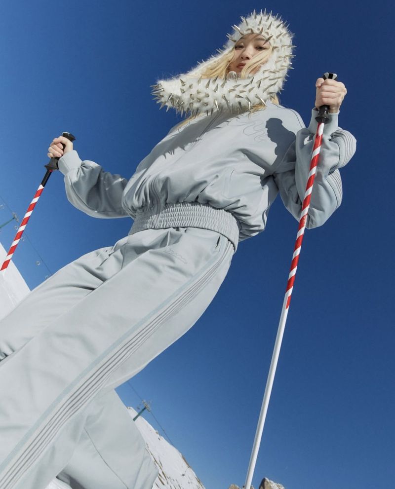 Low-angle shot of a female skier in a light grey outfit with a spiked hat holding ski poles against a clear blue sky and snowy ground.