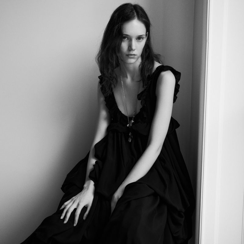 Black and white portrait of a woman with wavy brown hair, seated and wearing a black dress with ruffles, with a light-colored wall backdrop.