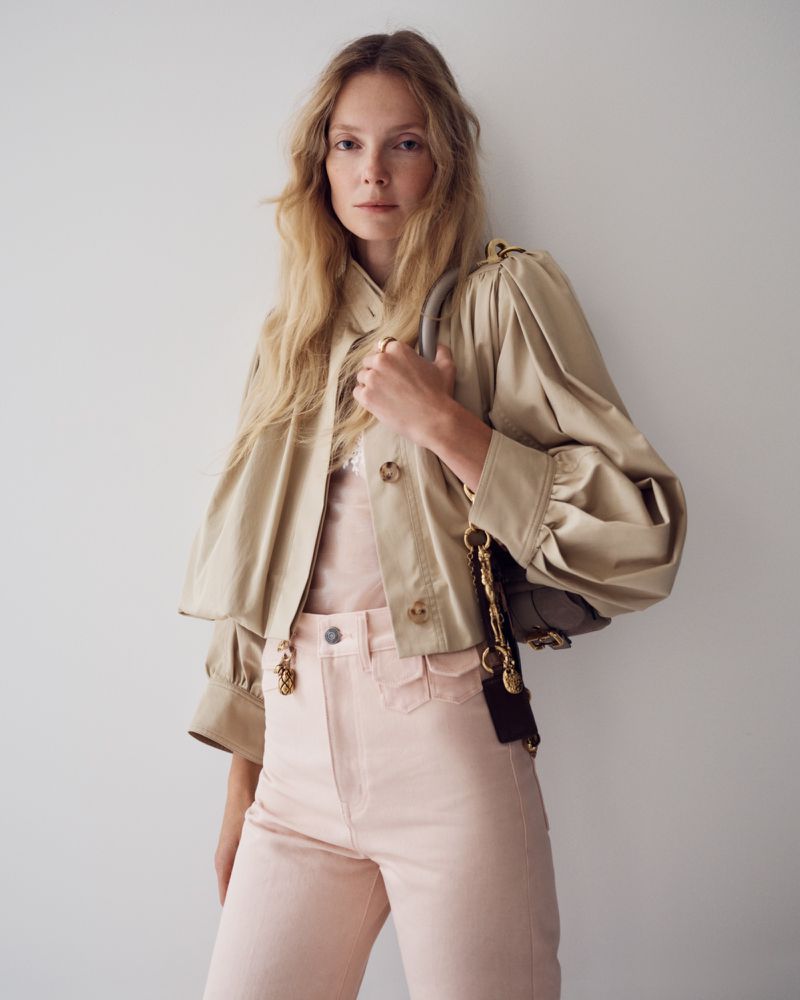 Blonde-haired woman in a beige jacket and pink pants with a purse, posing against a white background in a fashion shot.