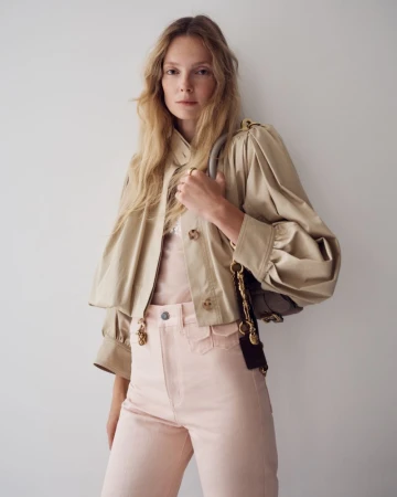 Blonde-haired woman in a beige jacket and pink pants with a purse, posing against a white background in a fashion shot. 