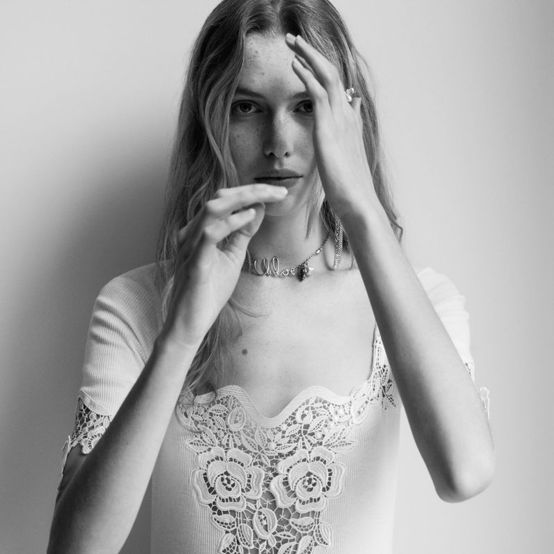 Monochrome portrait of a blonde woman with hands near her face wearing a lacy top and statement necklace in a photo studio setting.