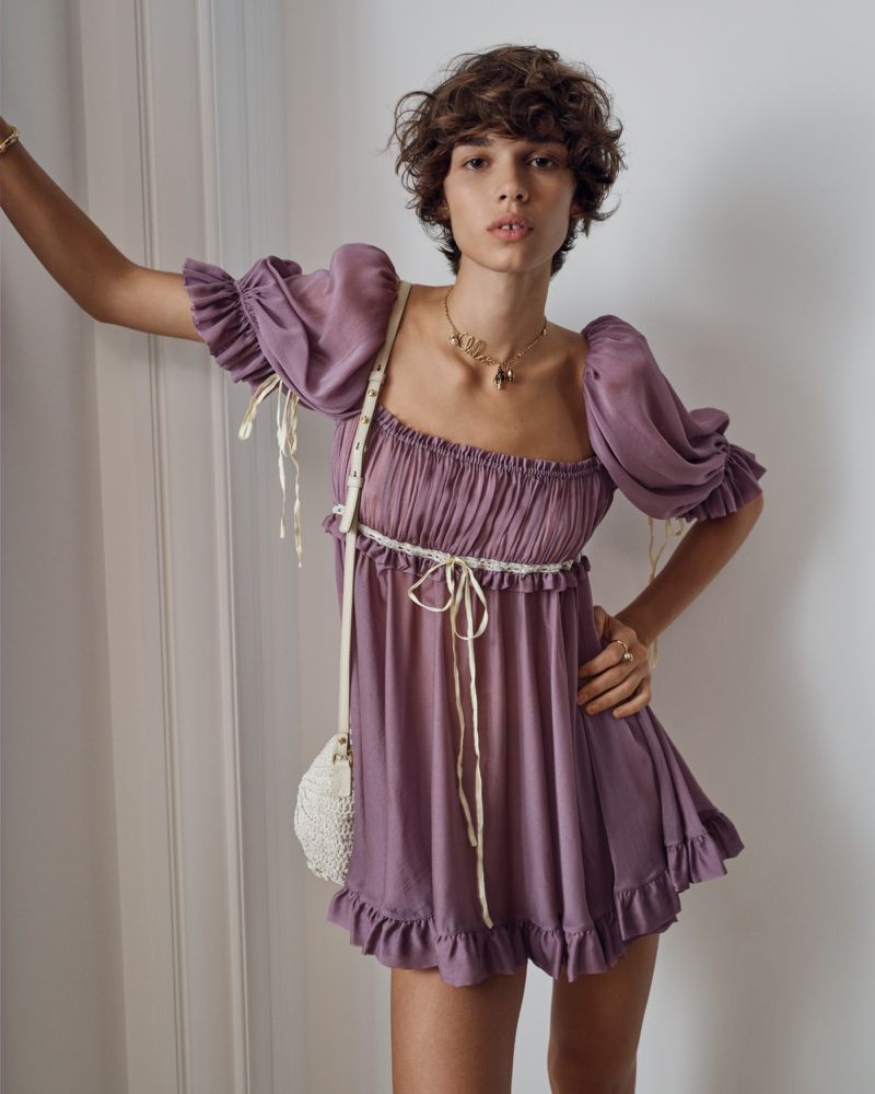 Woman in purple dress poses indoors by wall. Puff sleeves, cream lace trim, curly brown hair, necklace, and crossbody bag are visible.