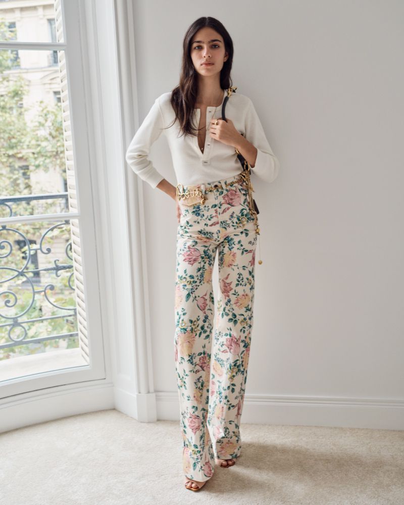 Elegant woman in a long-sleeved shirt and floral pants standing indoors by a bright window. She is holding a purse.