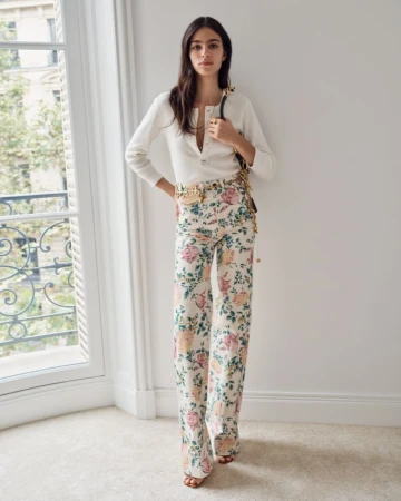 Model Loli Bahia poses in floral wide-leg trousers and a white henley top beside a tall French window.