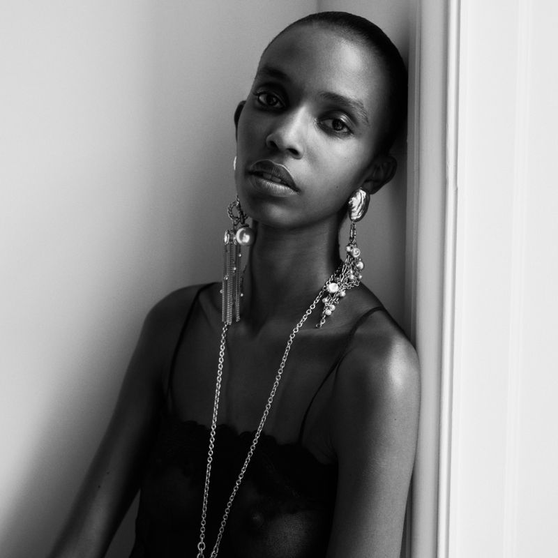 Monochrome portrait of a model wearing large dangling earrings and a long necklace against a plain wall.