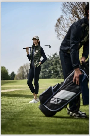 Two people on a golf course with one woman holding a golf club and wearing athletic wear. One golf bag is visible.