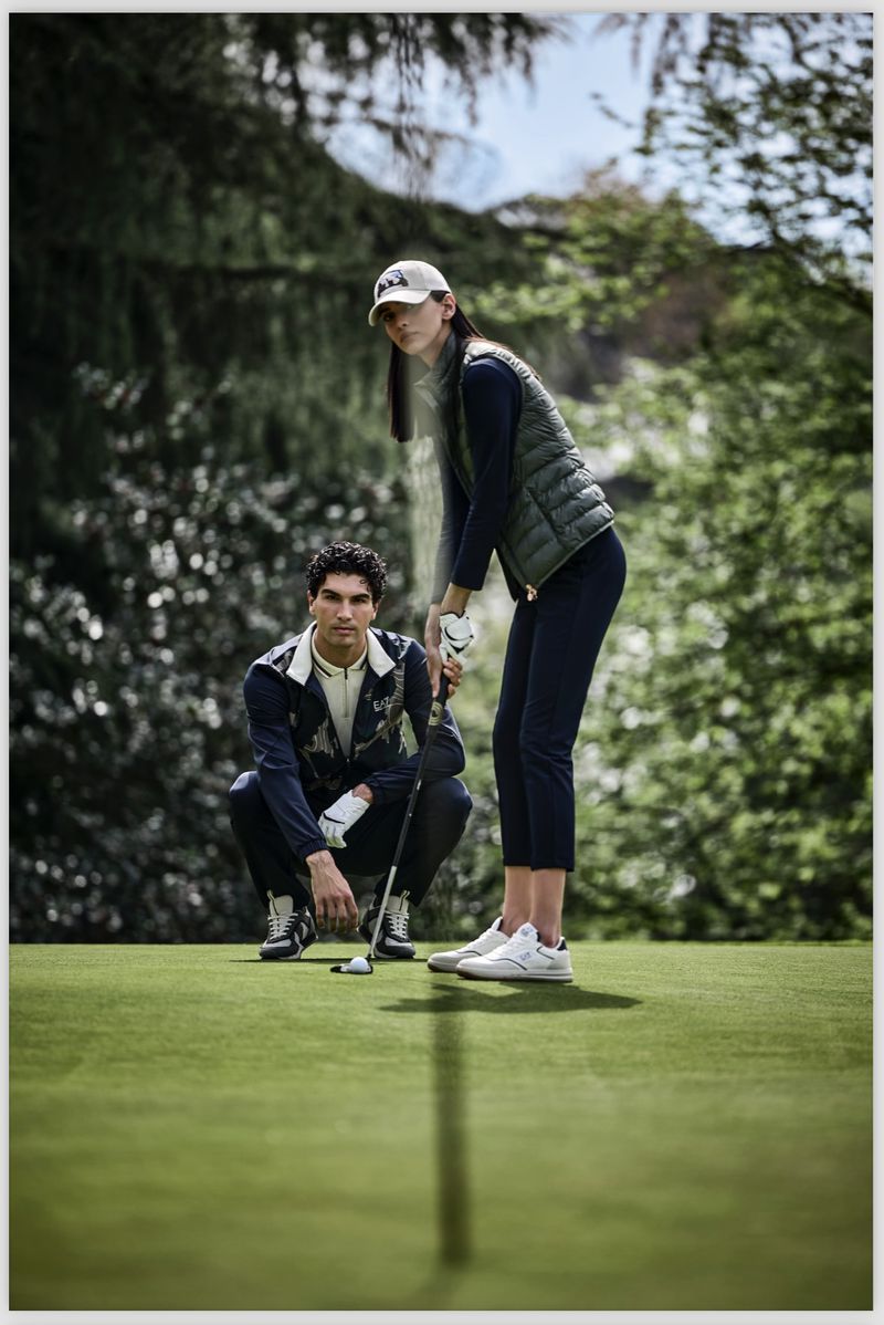 On a verdant golf course, a man and woman are poised for a game; she is about to swing as he watches in this outdoor shot.