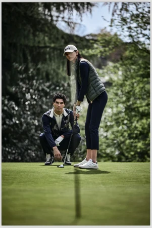 On a verdant golf course, a man and woman are poised for a game; she is about to swing as he watches in this outdoor shot.