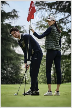 Two golfers on a green, man teeing off with golf club, woman holding a red flag, both wearing athletic apparel, daytime outdoor scene.