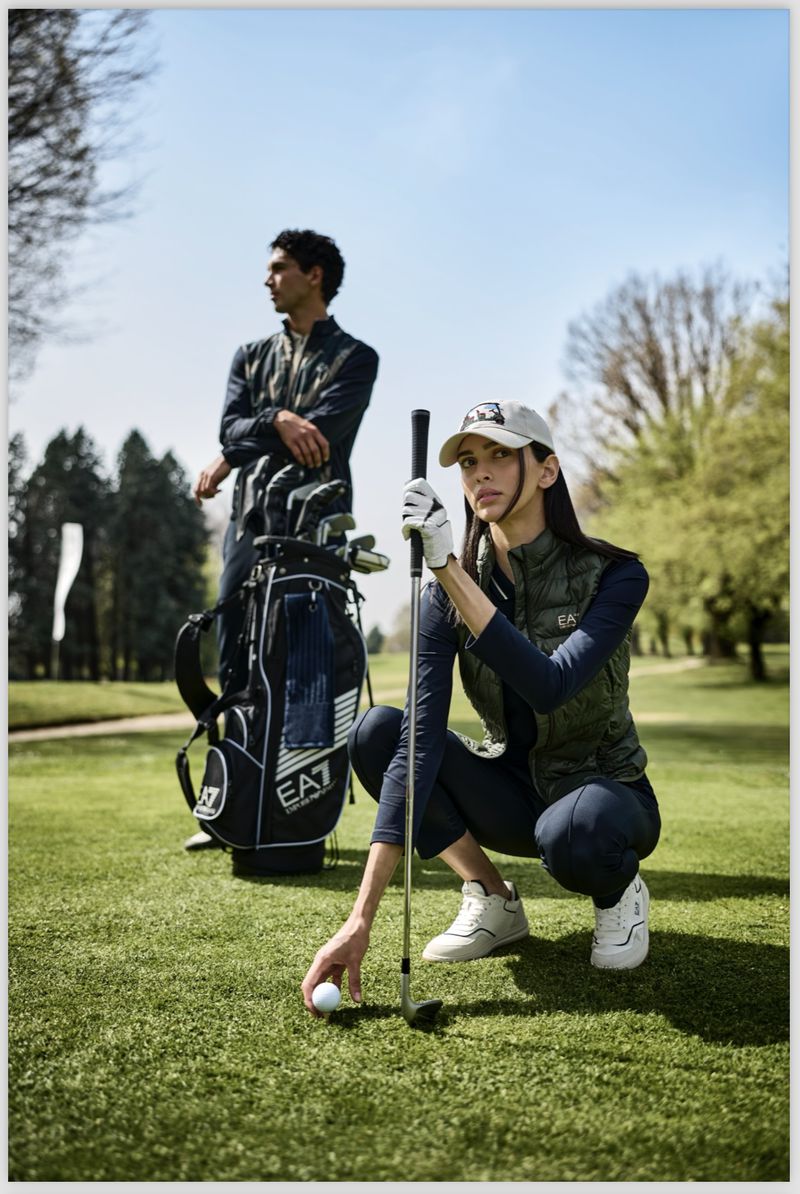 A fashionable couple on a sunny green golf course with golf clubs. The woman squats near her ball. Wearing sportswear, fashionable daytime scene.