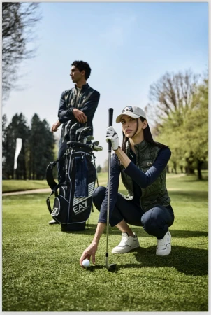 A fashionable couple on a sunny green golf course with golf clubs. The woman squats near her ball. Wearing sportswear, fashionable daytime scene.