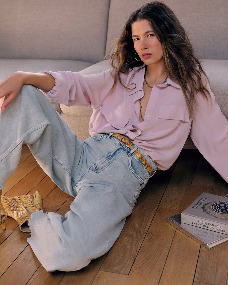 A woman in a lilac button-down and light wash jeans sits on a wood floor next to a beige couch with books in a casual pose.
