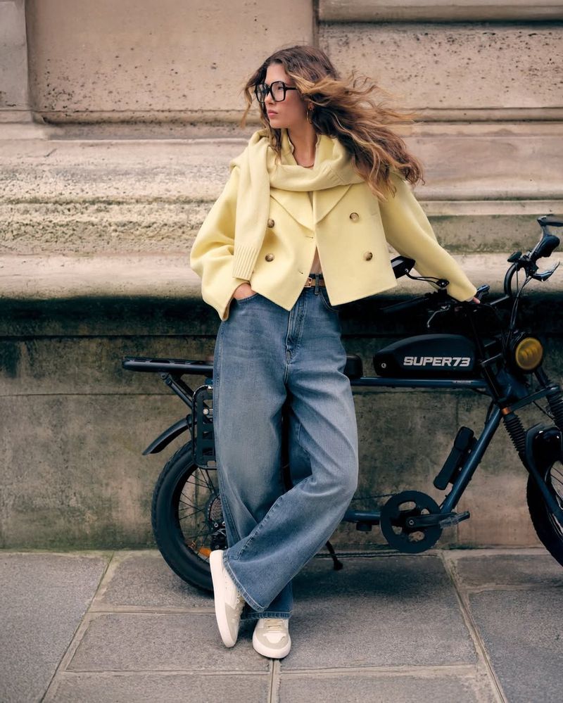 Adult woman in a yellow jacket, blue jeans, and white sneakers poses with an electric bicycle against a stone building in daylight.