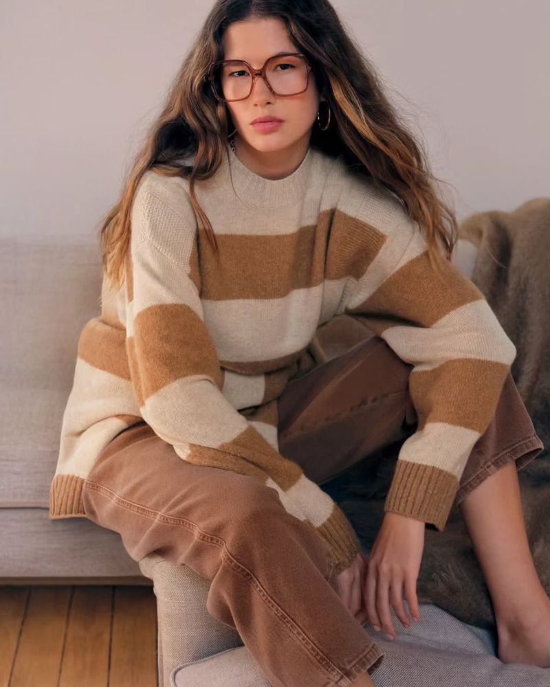 A woman with long brown hair and glasses is sitting indoors. She's wearing a striped beige and brown sweater and brown pants.