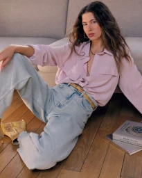 Model Meg Dmitruk sitting on a wood floor in a pale pink button-up shirt and wide-leg light wash denim jeans for Vanessa Bruno.